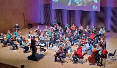 Ein mittelgro&szlig;es Jugendorchester probt in einem Saal unter einer Leinwand.