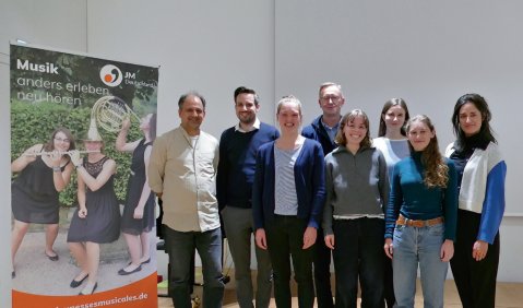 Eine Gruppe von Menschen steht mehrheitlich in allt&auml;glicher Kleidung neben einem Jeunesses-Musicales Aufsteller f&uuml;r ein Gruppenfoto in einem Probensaal zusammen.