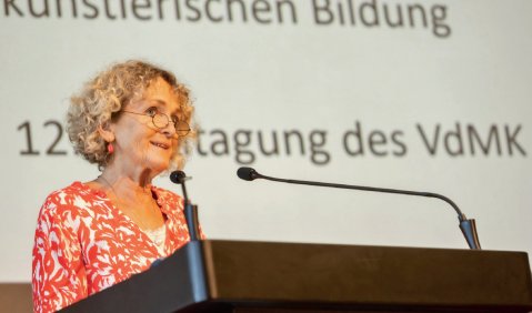 Eine Frau mit blonden Locken an einem Rednerpult. Im Hintergrund eine Projektsionsfl&auml;che mit Text.