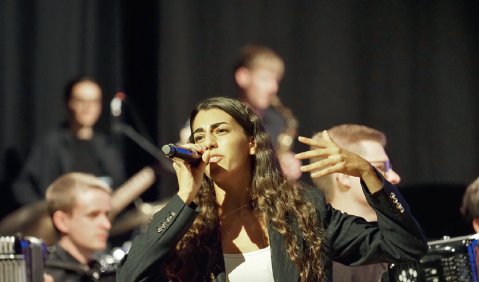 Eine junge erwachsene S&auml;ngerin mit braunen Locken in einem schwarzen Blazer vor einem Akkordeon-Orchester