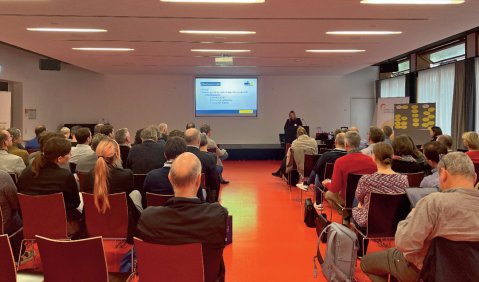 Ein mittlerer Seminarraum mit einer kleineren Leinwand, einer Rednerin und reichlich Publikum unter l&auml;nglichen Deckenleuchten.
