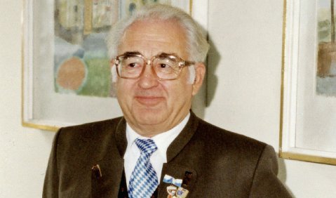 Ein &auml;lterer Mann mit wei&szlig;en Haaren und schwarzen Augenbrauen, Brille, leicht rundlichem Gesicht in einem dunkelbraunen Anzug, Weste, Taschenuhr-Kette, mehreren Anstecknadeln und einer blau-wei&szlig;en Krawatte