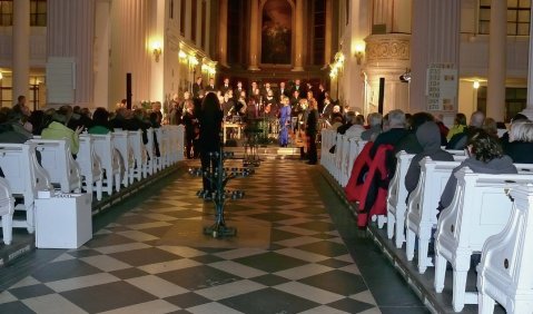 Wei&szlig;e Kirchenb&auml;nke in einer wei&szlig;en Kirche (in warmes Licht getaucht). Vor dem Altar stehen die Musiker:innen.