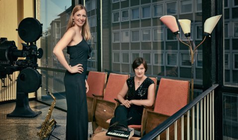 Zwei Frauen in dunklen Kleidern in einem Studio mit komplett verglaster R&uuml;ckwand. Eine sitzt auf einer ausgebauten Theaterstuhlreihe, die andere steht neben ihr. Ein Miniatur-Fl&uuml;gel steht auf einem Couchtisch zwischen ihnen, hinter ihnen ein Saxophon auf einem St&auml;nder.