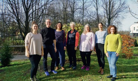 Sieben Personen in bunter Alltagskleidung in einem herbstlichen Garten.