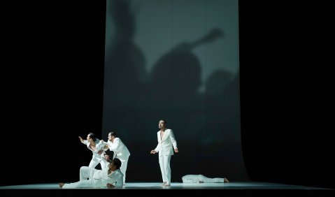 Fünf Rollen in ausschließlich weißer Kleidung werfen einen Schatten an das weiße Rechteck der sonst ausschließlich schwarzen und freien Bühnengestaltung