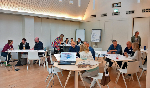 Ein weißer Seminarraum mit Holzboden und Tisch mit mehreren arbeitetenden Menschen daran.