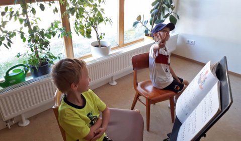 Zwei jungen sitzen auf kleinen Stühlen in einem Zimmer mit Zimmerpflanzen vor einem tiefen Fenster. Beide Jungen haben einen Blick in einen Notenständer.