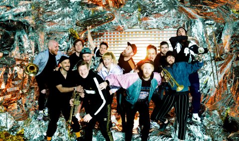 In einem Rahmen aus Erste-Hilfe-Folie posiert die 13-Köpfige Band mit ohne Blasinstrumenten in einem wilden Getummel.