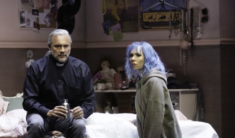 Ein Pastor auf einem Bett in einem Teenager-Zimmer, die Teenagerin mit blauen Haaren kniet neben ihm auf dem Boden, beide gucken suchend ins Publikum.