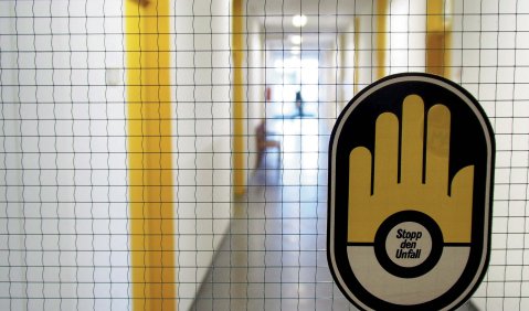 Ein weißer Flur mit gelben Türrahmen und grauem Boden durch eine hauchdünn quadratisch gegitterte Glasscheibe fotografiert. An der Glasscheibe klebt ein Aufkleber mit einem Handsymbol in denssen Handfläsche steht: "Stopp den Unfall"