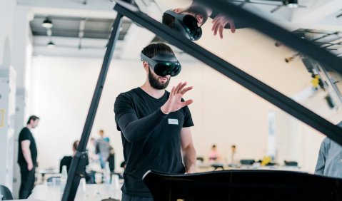 Ein jüngerer Mann steht mit einer VR-Brille vor den Augen (also einer etwas vorstehenden schwarzen gläsernen großen und dicken Brille) vor einem offenen Flügel und streckt di9e Hand aus. Das ganze ist in einer laborähnlichen weißen Halle.