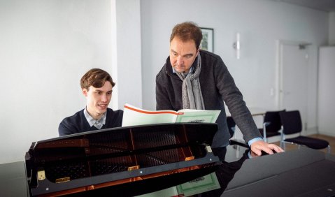 Ein jüngerer Mann mit kurzen leicht gewellten braunen Haaren sitzt an einem Flügel, ein ihm gegenüber älterer Mann steht, auf den Flügel gestützt, neben ihm: Sie schauen zusammen in die auf dem Instrument stehenden Noten.