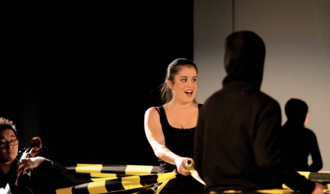 Konzertperformance oder szenisches Konzert: Eine Frau steht in einem von schwarz-gelbem Absperrband eingerahmten Bereich, links von ihr ist ein Teil eines Cellisten zu sehen. Vor ihr ist eine Gestalt die nur als schwarzer Schatten wahrgenommen werden kann.