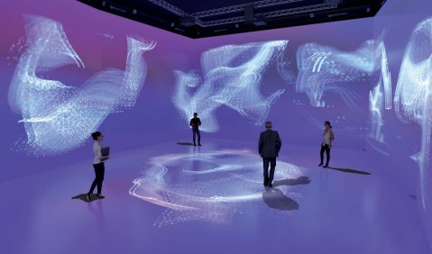 4 Personen in einem bühnenähnlichen Raum mit Projektionsflächen als Boden und Wänden. Sie stehen in der Mitte des Raumes und in ihrer Mitte, wie auch an den Wänden sind helle geometrische Projektionen. Die Flächen sonst sind eher Blau-Lila gehalten.
