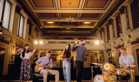 Mehrere junge Menschen stehen für das Foto zusammen und bedienen unterschiedlichste Instrumente (von Drumset und Gitarre bis Flöte) in einem klassizistischen Saal oder Theater.