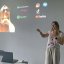 Eine junge blonde Frau mit lockeren weißen Klamotten hält einen Vortrag vor einer weißen Wand, an die diverse Logos von Social-Media- und Musik-Streaming-Anbietern anprojiziert werden.