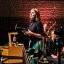 Die Frau im jungen mittleren Alter moderiert vorm Orchester stehend. Die aus vielen unregelmäßgen Akustik-Panelen bestehende Elbphilharmonie-Rückwand ist rost-rot angeschienen.