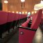 Rote Polster-Theaterstühle mit schwarzem Gestellt auf schwarzem Boden. Im Hintergrund eine cremefarbene Wand mit reichlich Leuchten. An der fotografierten Stuhlreihe eine Nummer: 9.